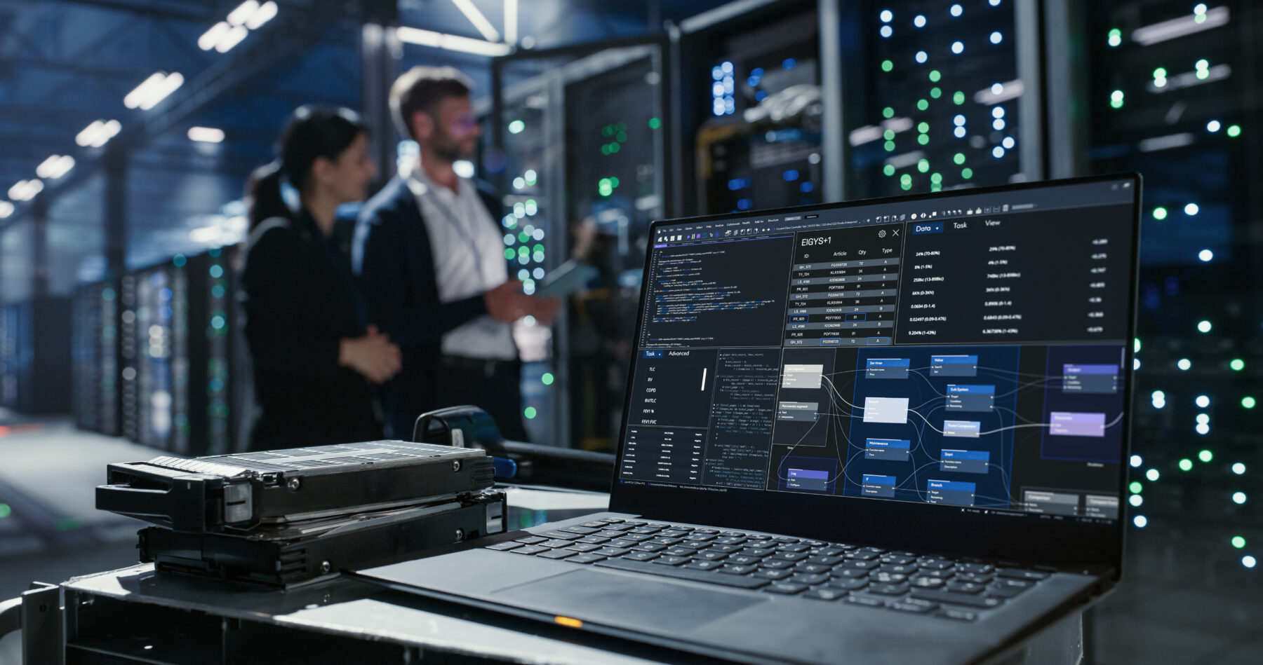 Laptop sits on a cart beside hard drives in data center. Two people discuss network infrastructure and server maintenance among server racks and mainframes.